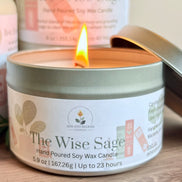 Candle in a tin labeled 'The Wise Sage' with a lit flame, surrounded by greenery on a wooden surface.