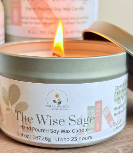 Candle in a tin labeled 'The Wise Sage' with a lit flame, surrounded by greenery on a wooden surface.