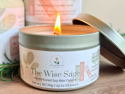 Candle in a tin labeled 'The Wise Sage' with a lit flame, surrounded by greenery on a wooden surface.