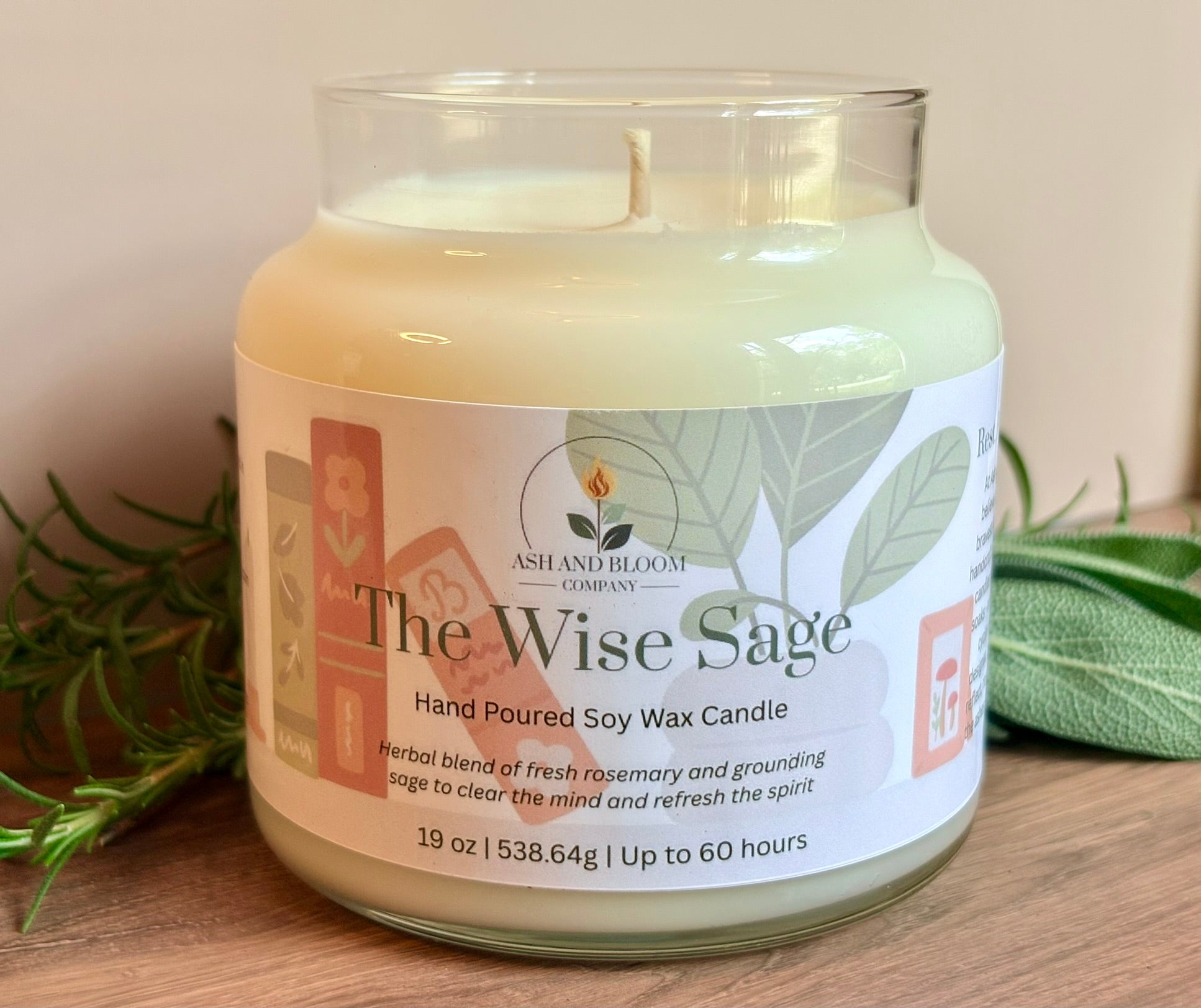 Candle labeled 'The Wise Sage' from Ash & Bloom Company on a wooden surface with greenery.