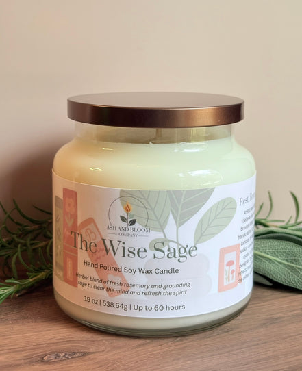Rosemary and sage candle jar labeled 'The Wise Sage' on a wooden surface with greenery in the background