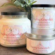 Three 'Saging Gracefully' candles of different sizes on a wooden surface with a plant in the background.