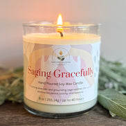 Lavender and sage candle with a label reading 'Saging Gracefully' on a wooden surface with greenery.