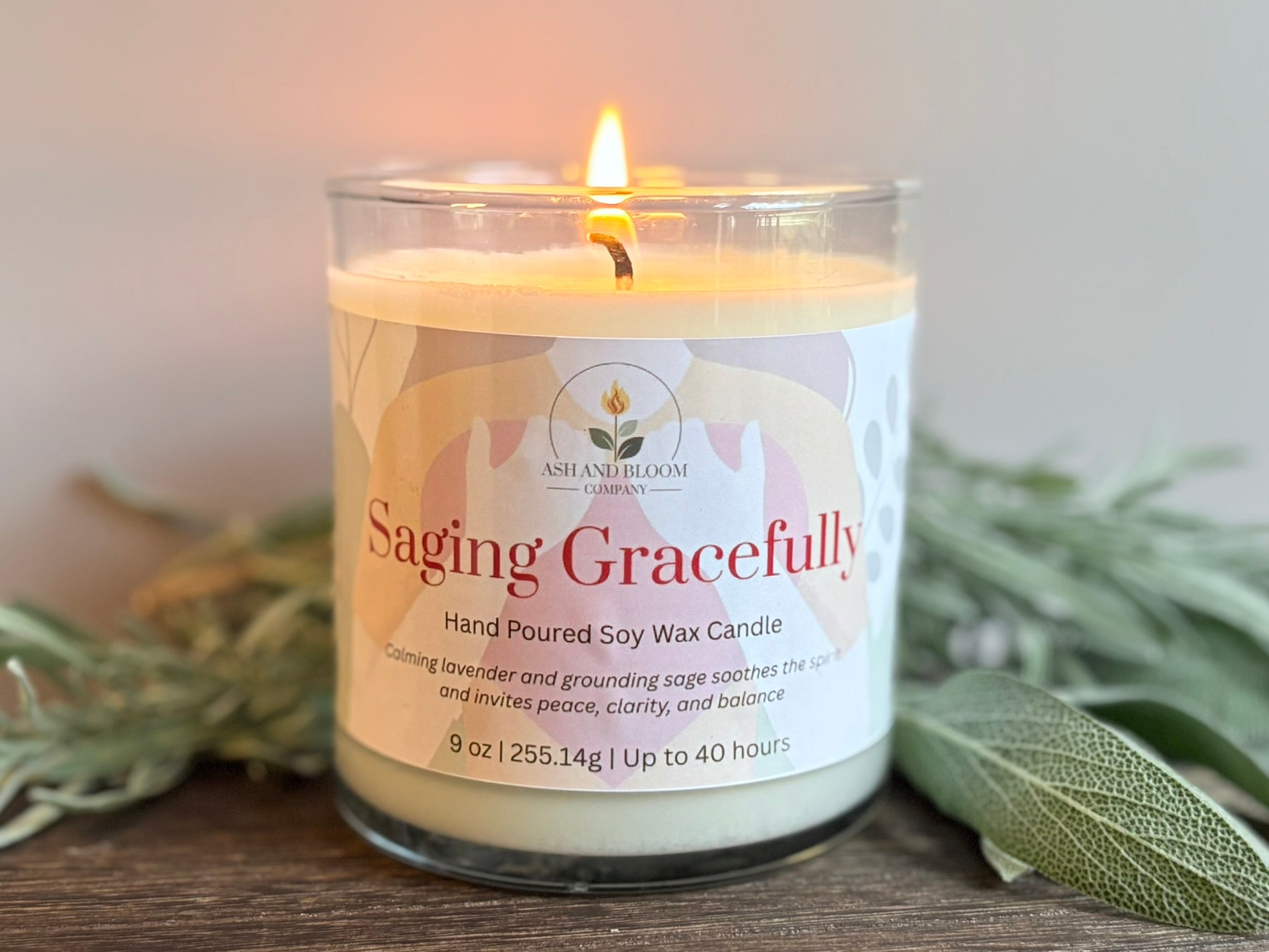 Lavender and sage candle with a label reading 'Saging Gracefully' on a wooden surface with greenery.