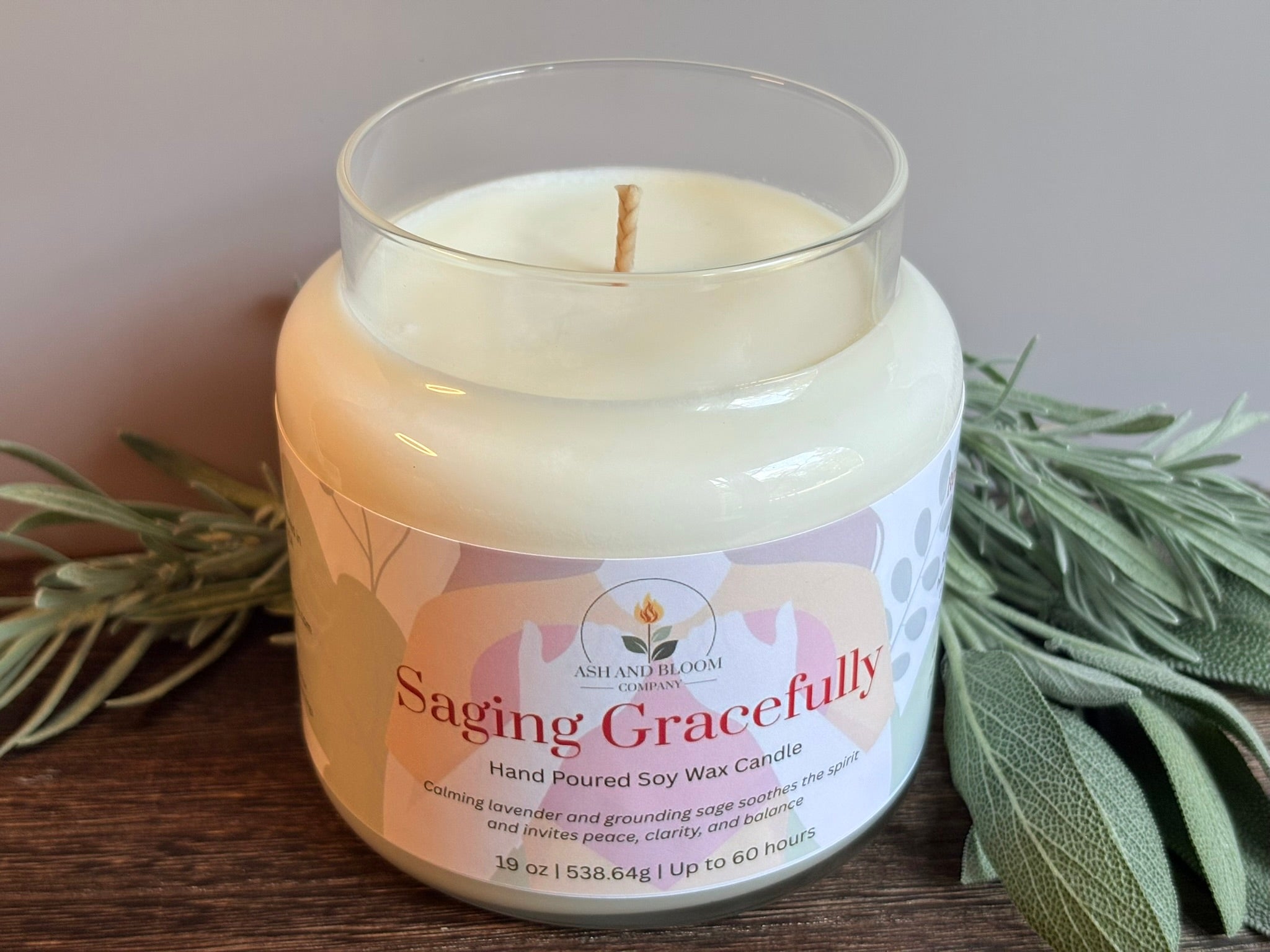 Candle in a glass jar with a label reading 'Saging Gracefully' on a wooden surface with sage leaves.