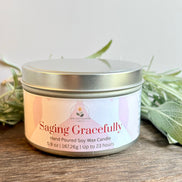 Lavender and Sage Saging Gracefully candle in a metal tin with a white label on a wooden surface with green leaves.