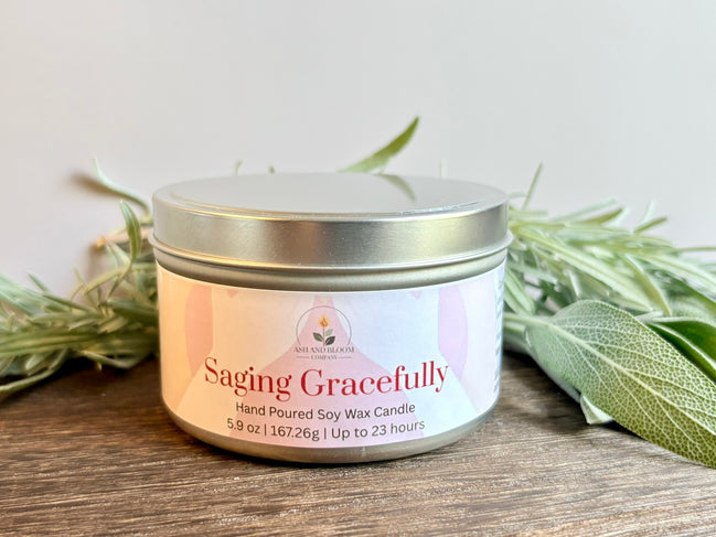 Lavender and Sage Saging Gracefully candle in a metal tin with a white label on a wooden surface with green leaves.
