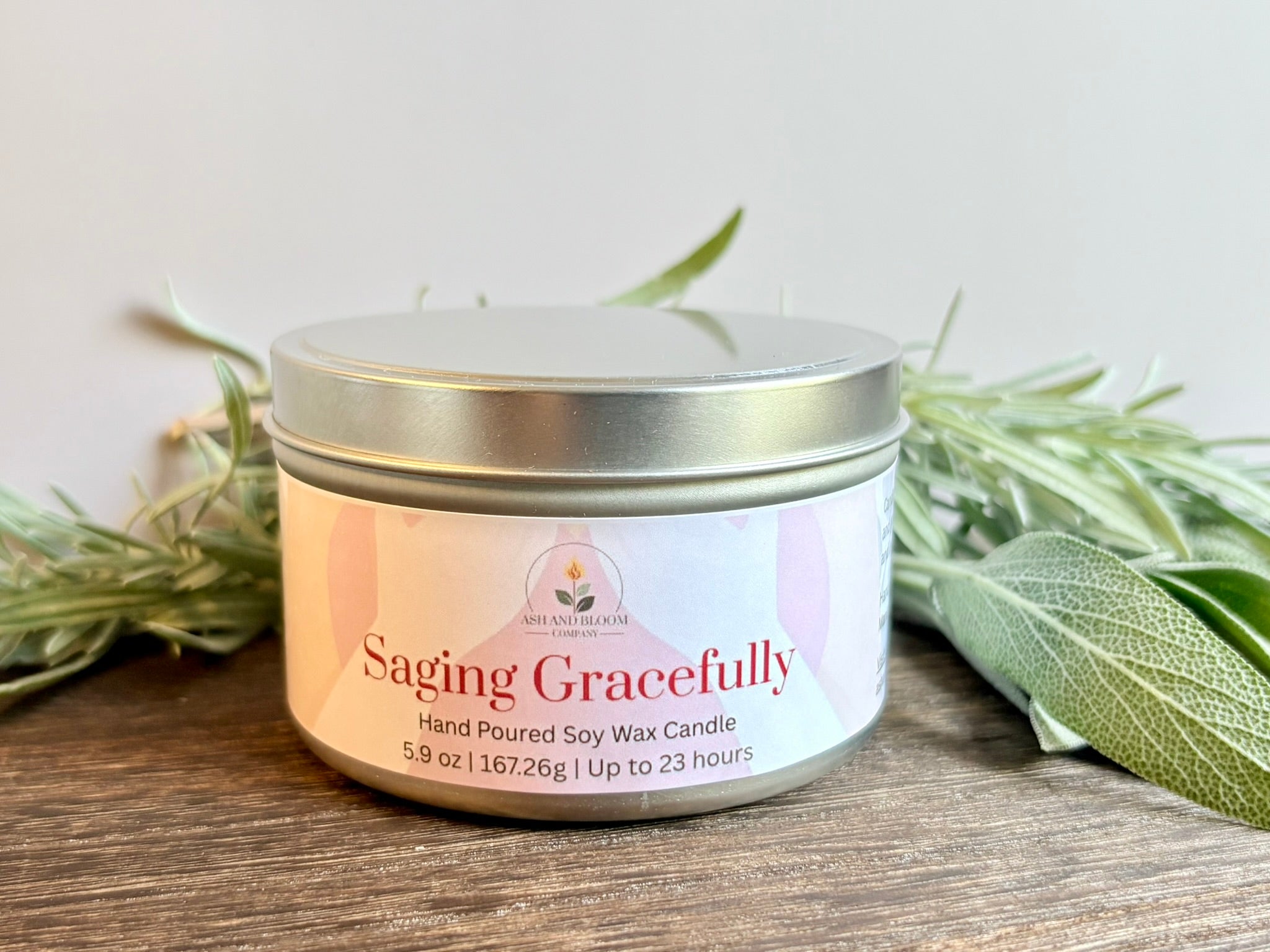 Lavender and Sage Saging Gracefully candle in a metal tin with a white label on a wooden surface with green leaves.
