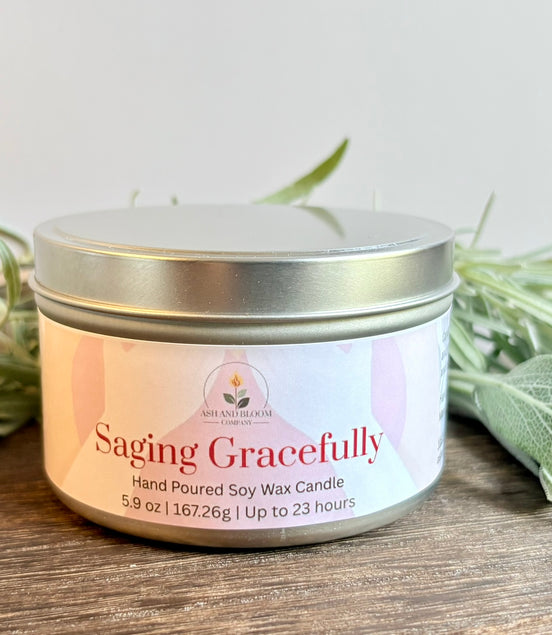Lavender and Sage Saging Gracefully candle in a metal tin with a white label on a wooden surface with green leaves.