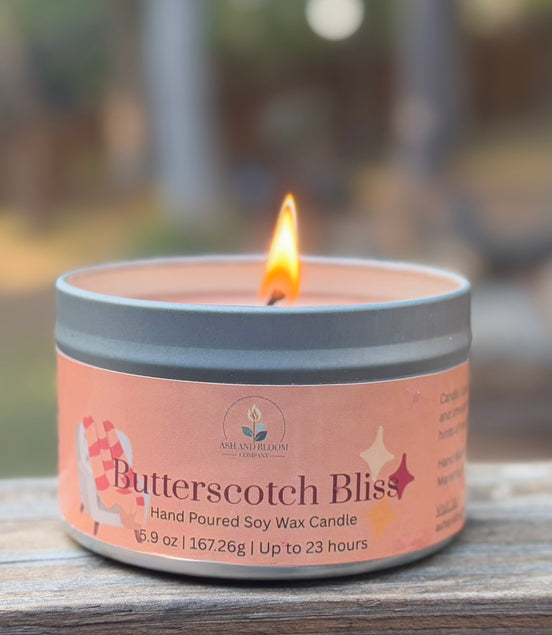 Candle labeled 'Butterscotch Bliss' on a wooden surface with a blurred outdoor background