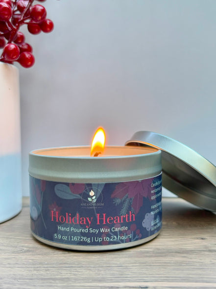 Candle in a tin labeled 'Holiday Hearth' with a lit candle inside, on a wooden surface.