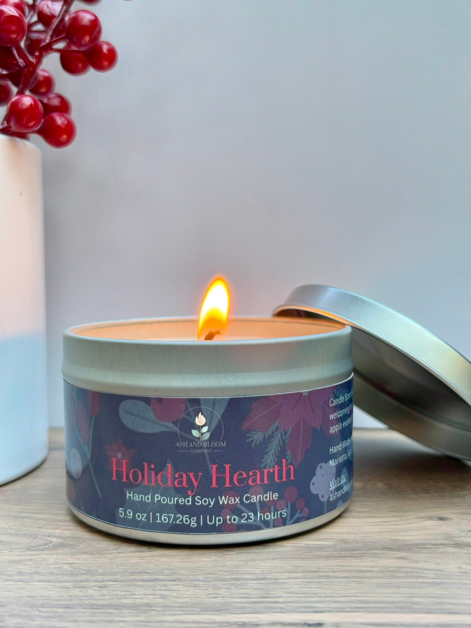 Candle in a tin labeled 'Holiday Hearth' with a lit candle inside, on a wooden surface.