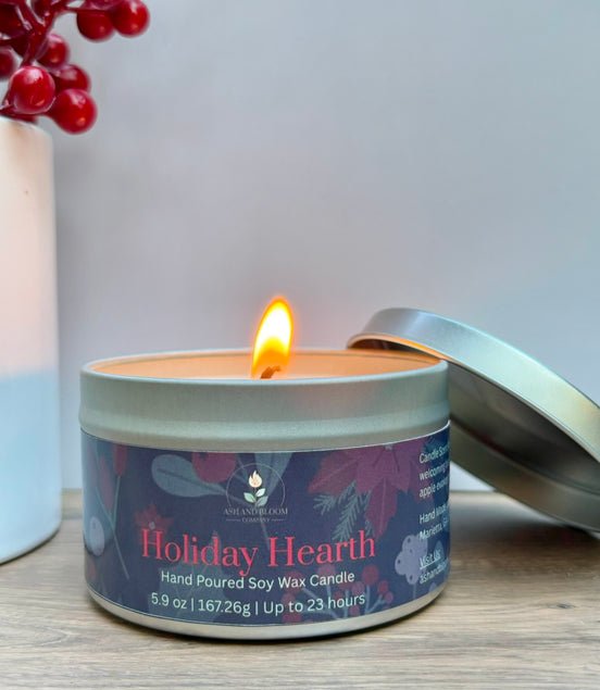 Candle in a tin labeled 'Holiday Hearth' with a lit candle inside, on a wooden surface.
