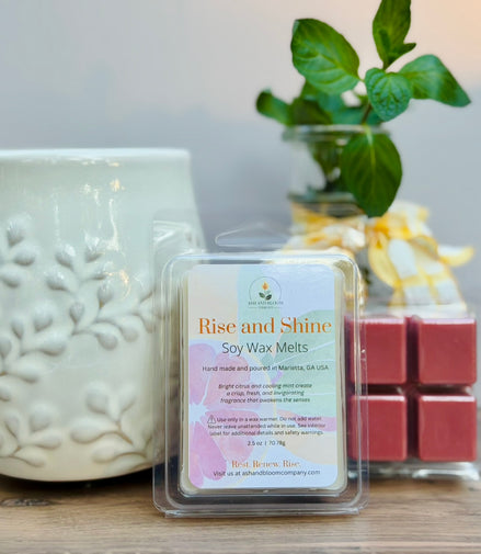 Pack of 'Rise and Shine' citrus and mint soy wax melts next to a textured ceramic wax melt container on a wooden surface.