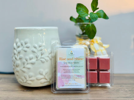 Pack of 'Rise and Shine' citrus and mint soy wax melts next to a textured ceramic wax melt container on a wooden surface.