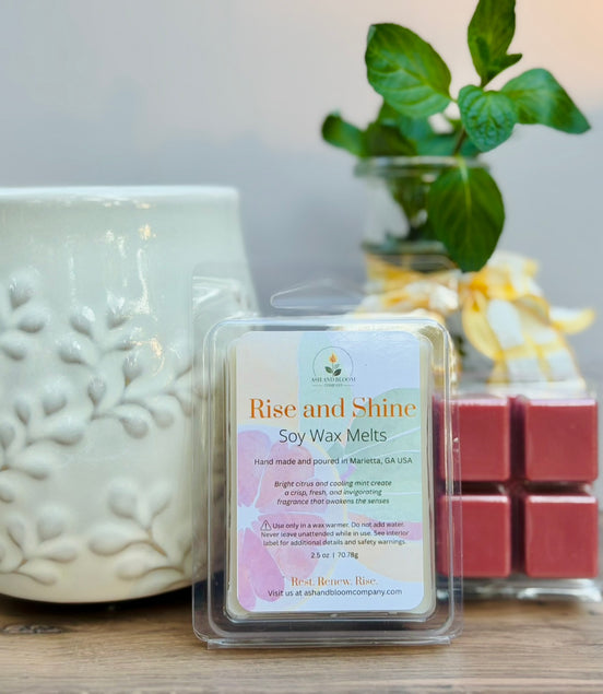 Pack of 'Rise and Shine' citrus and mint soy wax melts next to a textured ceramic wax melt container on a wooden surface.