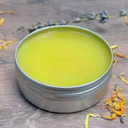 Metal tin with greenish-yellow salve on a wooden surface with dried flowers.