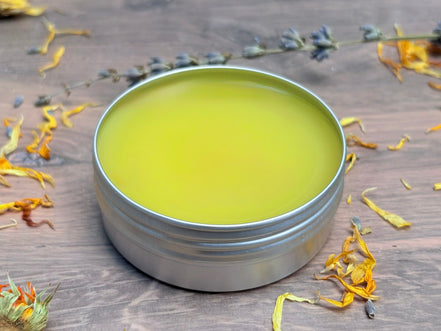 Metal tin with greenish-yellow salve on a wooden surface with dried flowers.