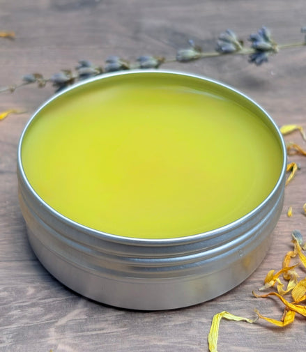 Metal tin with greenish-yellow salve on a wooden surface with dried flowers.