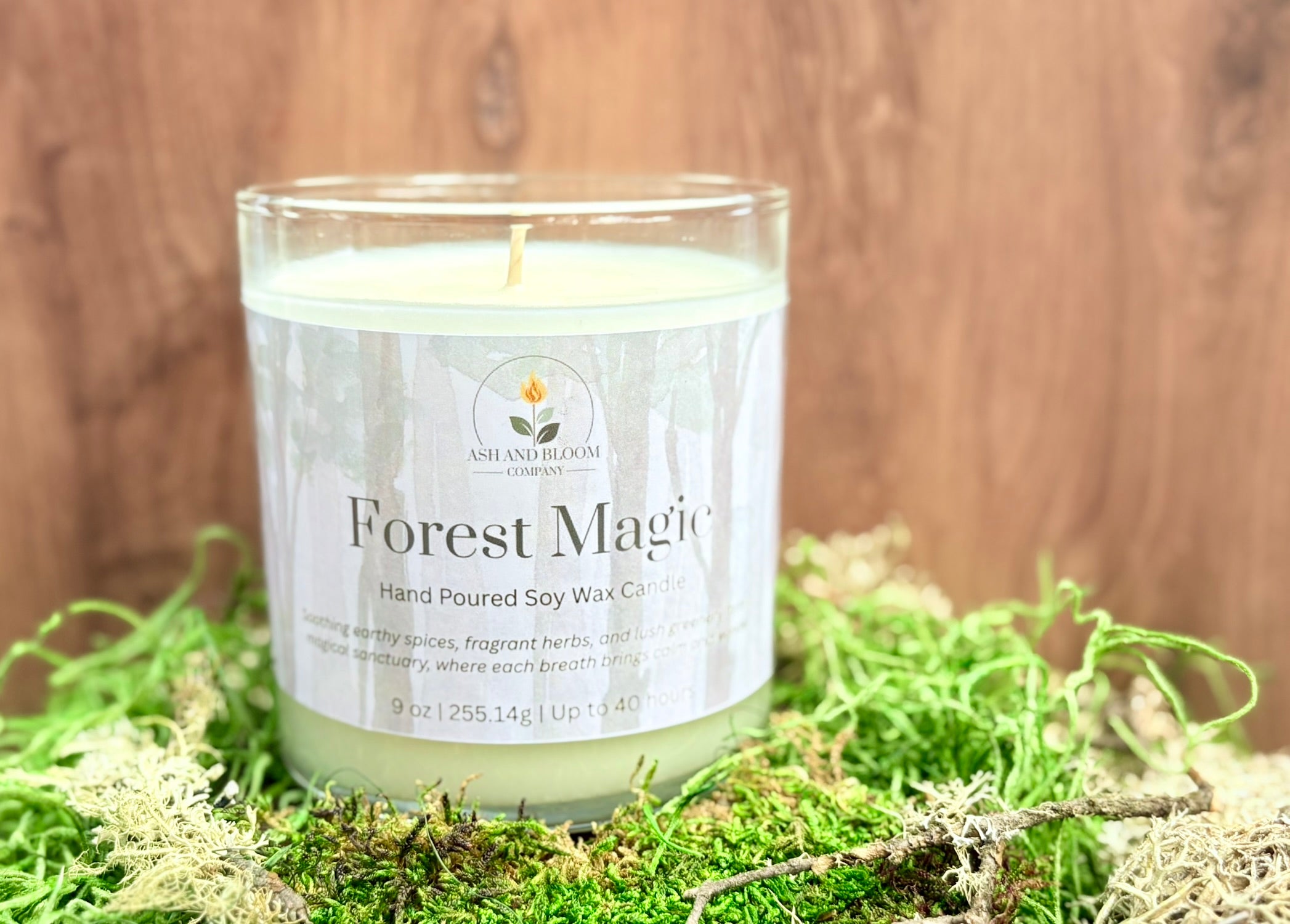 Candle labeled 'Forest Magic' on a bed of green moss with a wooden background