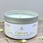 White tea and jasmine scented candle in a tin labeled 'Clari-tea' on a wooden surface with tea leaves.