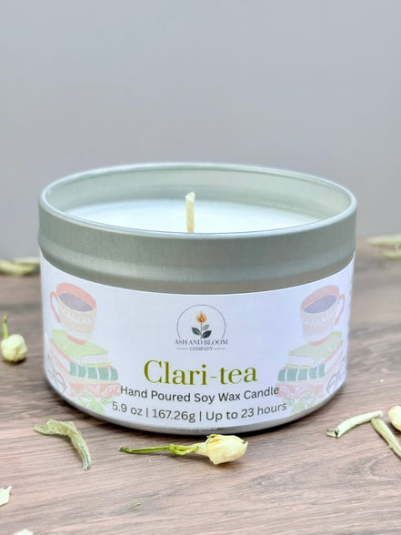 White tea and jasmine scented candle in a tin labeled 'Clari-tea' on a wooden surface with tea leaves.