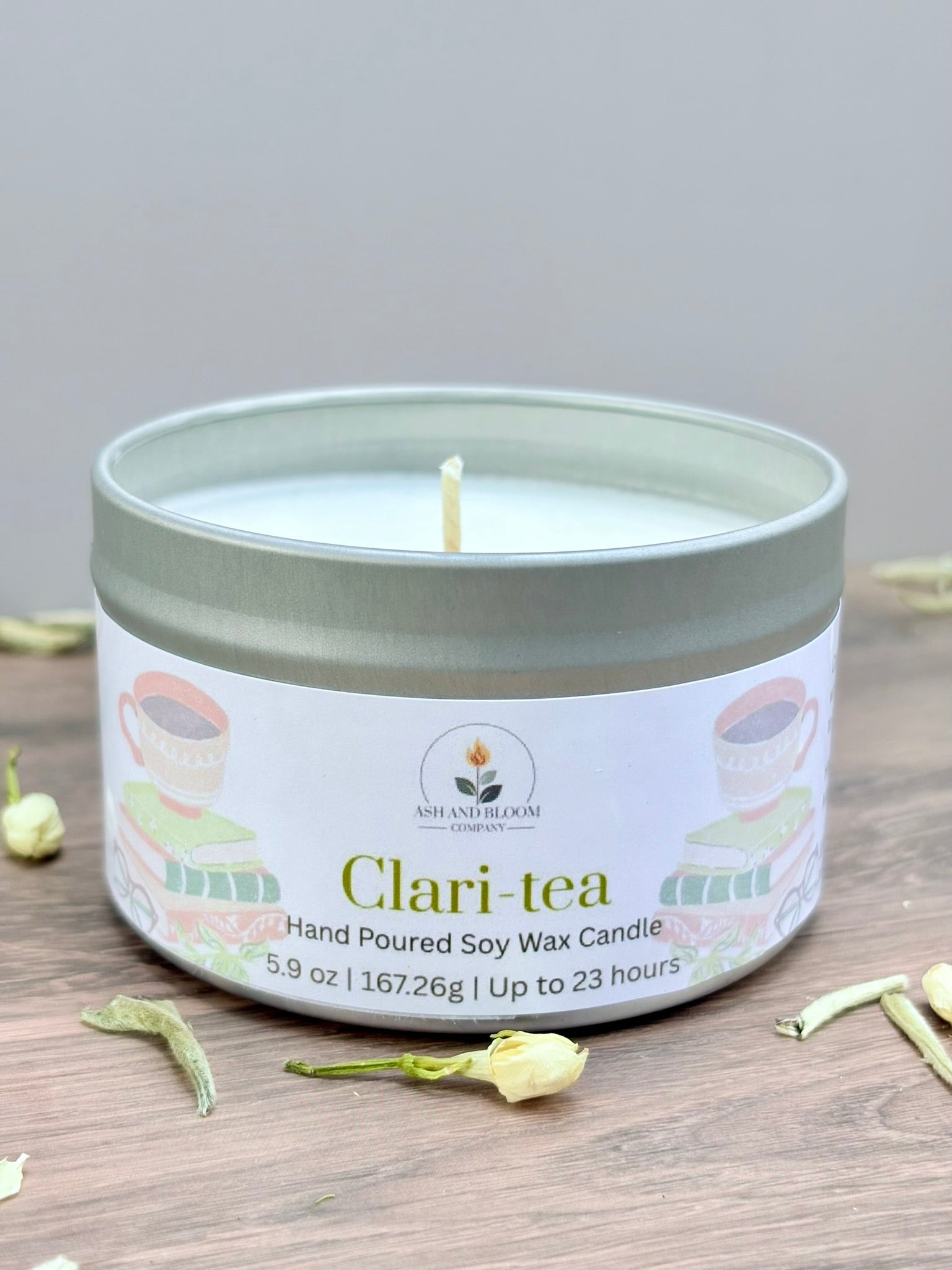 White tea and jasmine scented candle in a tin labeled 'Clari-tea' on a wooden surface with tea leaves.