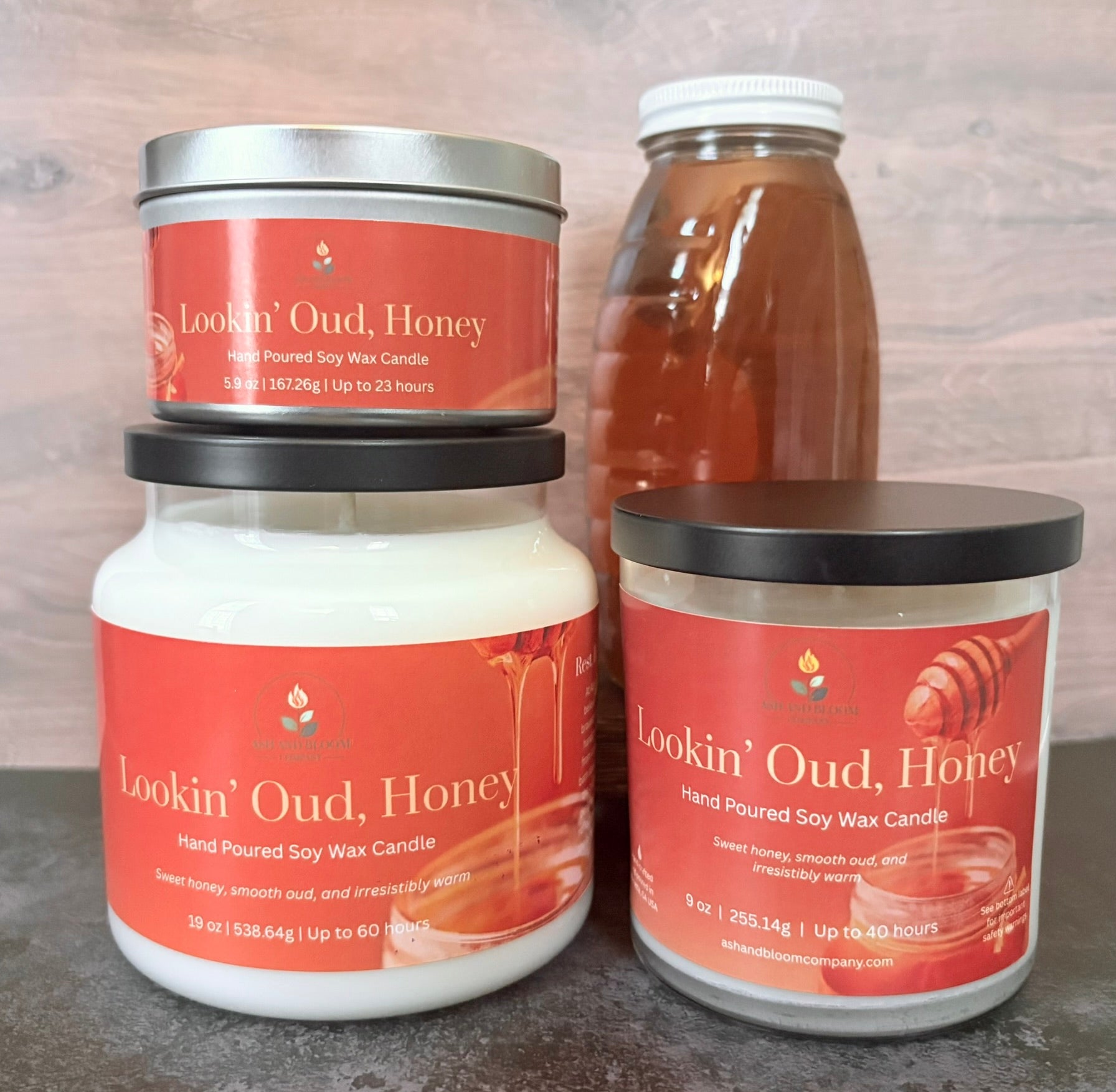 Three 'Lookin' Oud, Honey' candles of different sizes on a wooden surface with a bottle of honey in the background