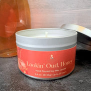 Candle in a tin labeled 'Lookin' Oud, Honey' on a textured surface.