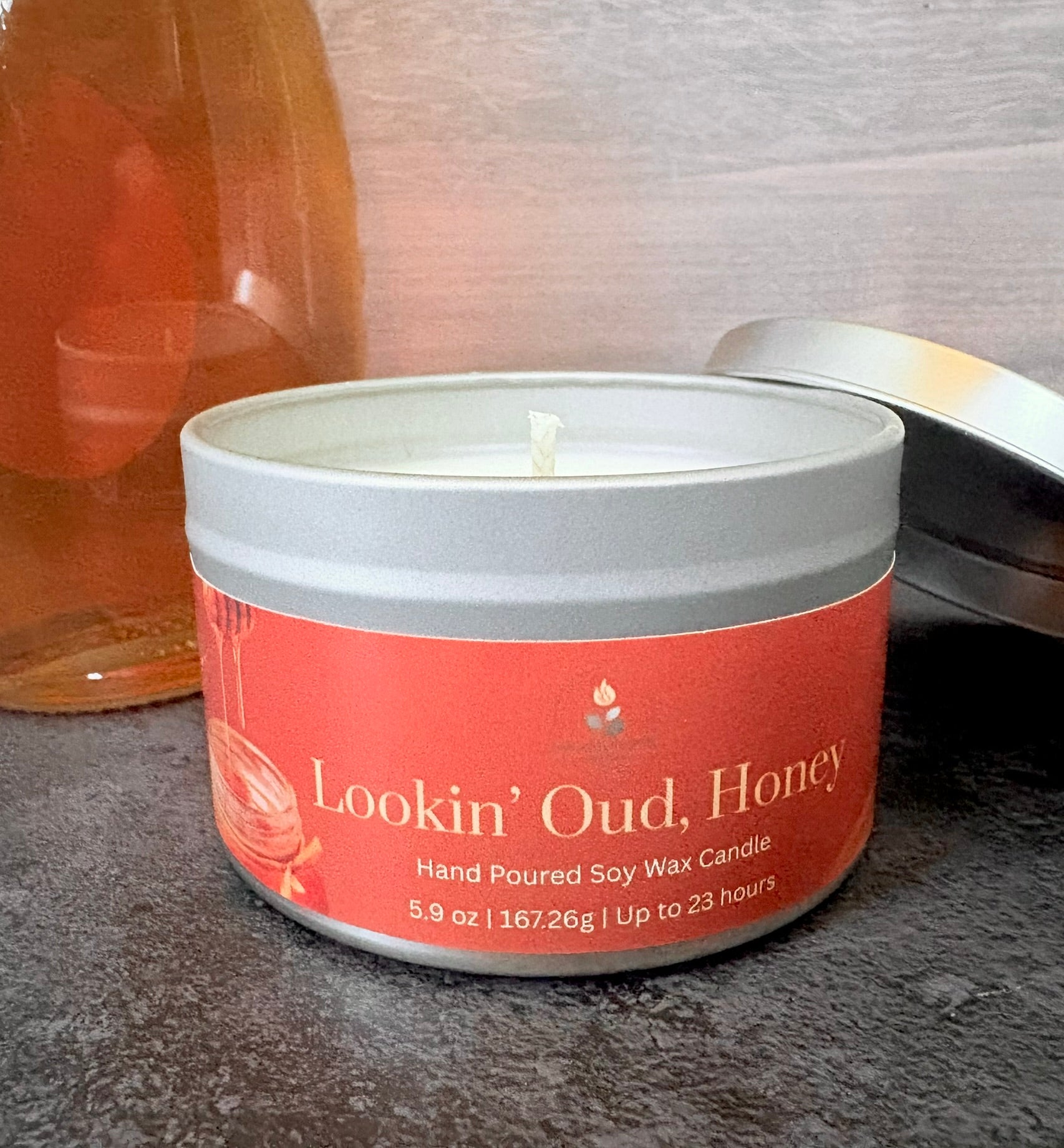 Candle in a tin labeled 'Lookin' Oud, Honey' on a textured surface.